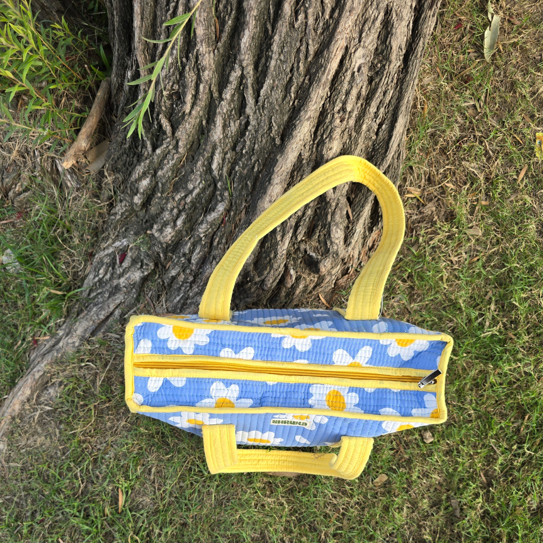 A blue and white daisy-patterned fabric bag with bright yellow handles and zipper trim, photographed outdoors against a tree trunk and grass.